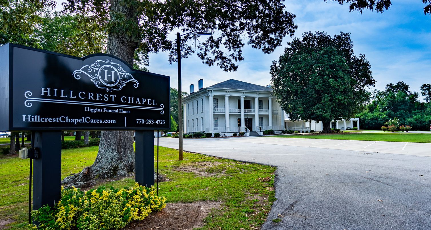 A black Hillcrest Chapel sign in the foreground, with a white, two-story antebellum-style building in the background.