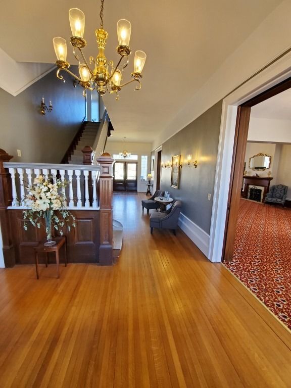 A hallway with polished hardwood floors, a wooden staircase, a chandelier, and a wide doorway into a carpeted room.