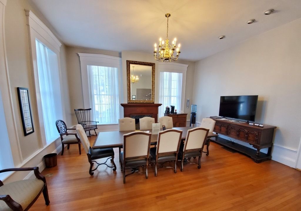 A dining room with a long wooden table, six upholstered chairs, a fireplace, a mirror, and a television on a side table.