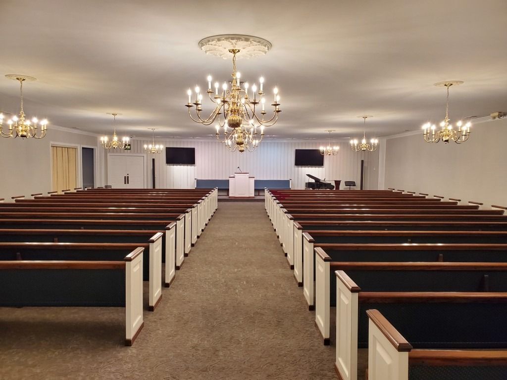 An empty church sanctuary with rows of dark wood pews facing a central podium, lit by several ornate chandeliers.