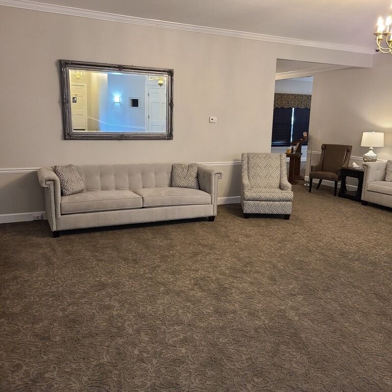 A living room with a neutral-toned sofa and armchair, a rectangular mirror on the wall, and carpeted flooring.