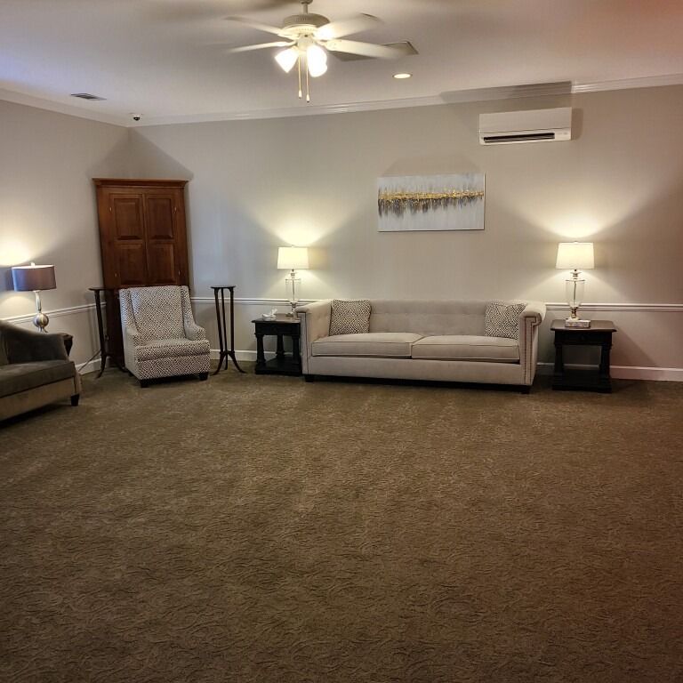 A neutral-toned living room featuring a beige sofa, patterned armchair, side tables, lamps, and a ceiling fan.
