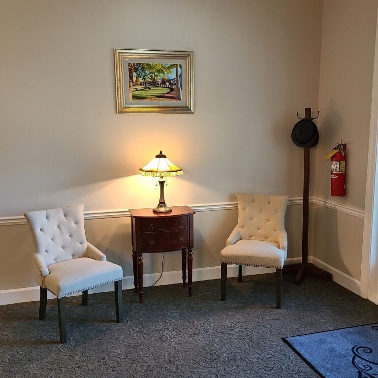 Two tufted beige chairs, a dark wood side table with a lit lamp, a framed picture, and a coat rack with a hat.