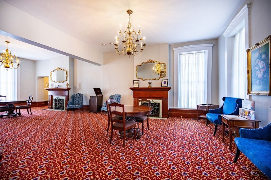 A formal, well-lit parlor with red patterned carpeting, two fireplaces with mirrors, chandeliers, and blue armchairs.