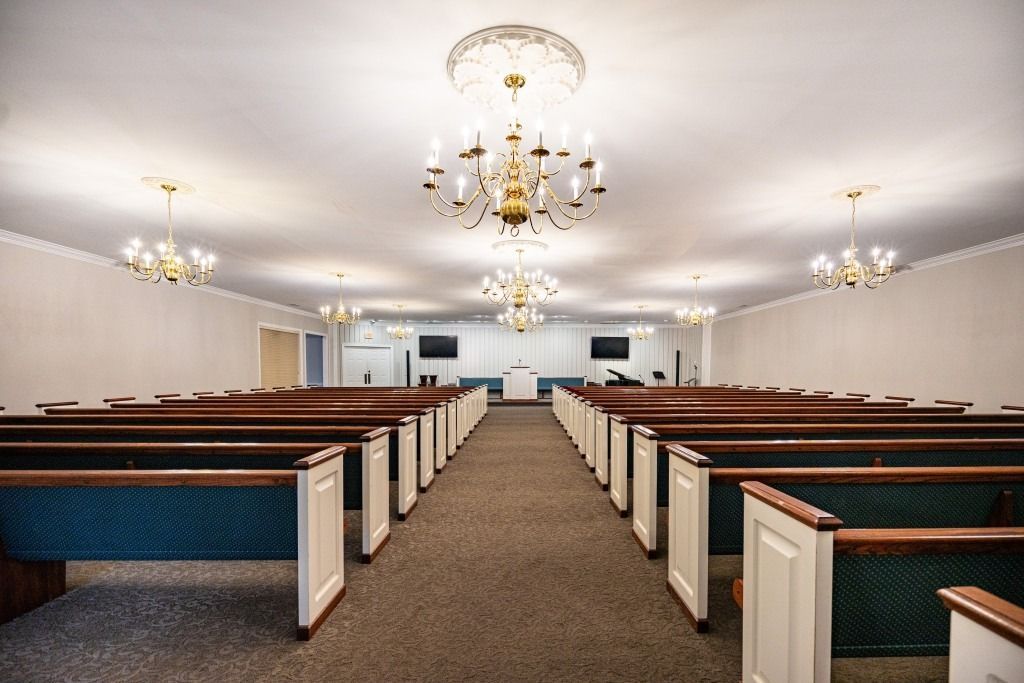 Interior of a quiet, empty chapel with rows of wooden pews, carpeted floors, and multiple elegant chandeliers.