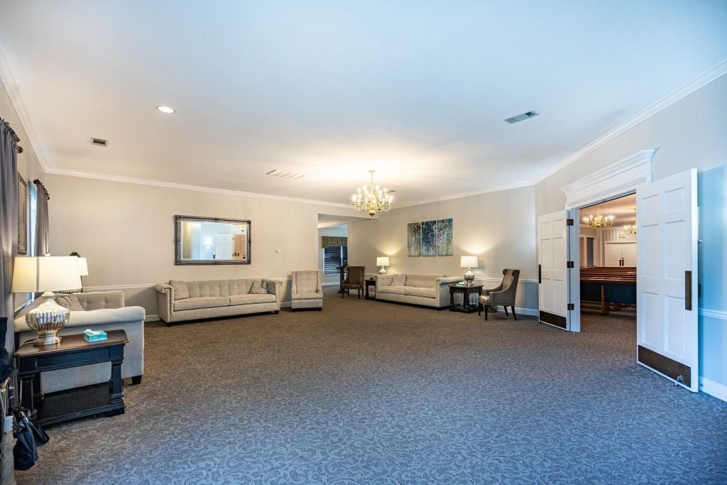 A spacious, neutrally decorated room with sofas, armchairs, two crystal chandeliers, and gray carpeting.