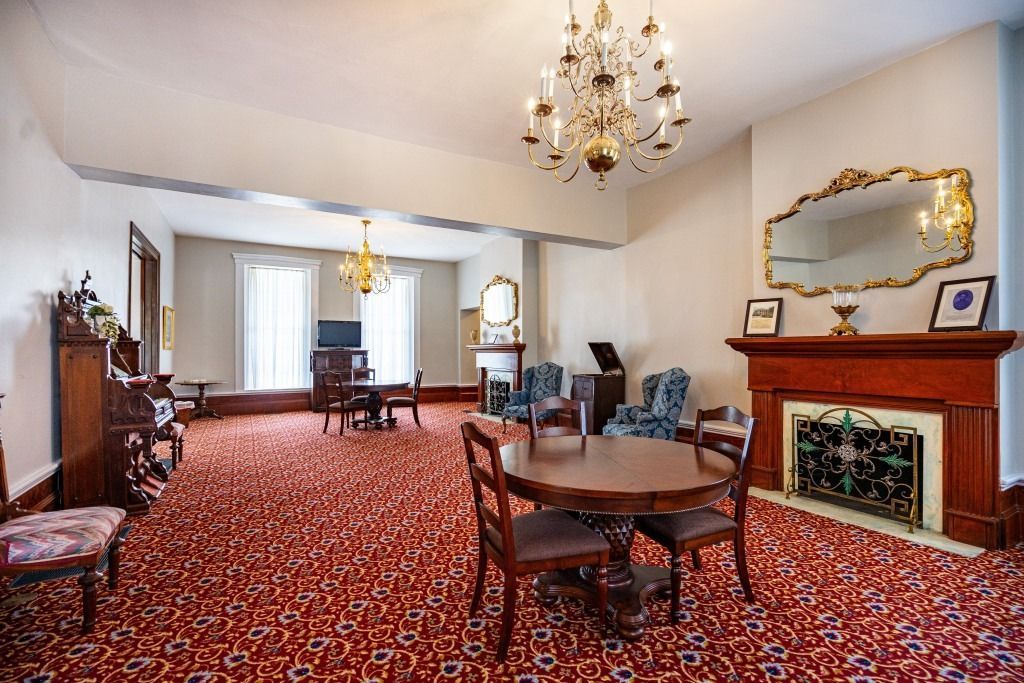 A spacious, formal room with red patterned carpet, chandeliers, a fireplace, and several small seating areas.
