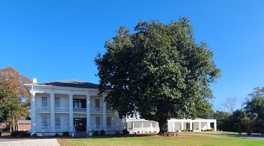 A large, white, two-story colonial-style house with pillars stands next to a massive green tree under a clear blue sky.
