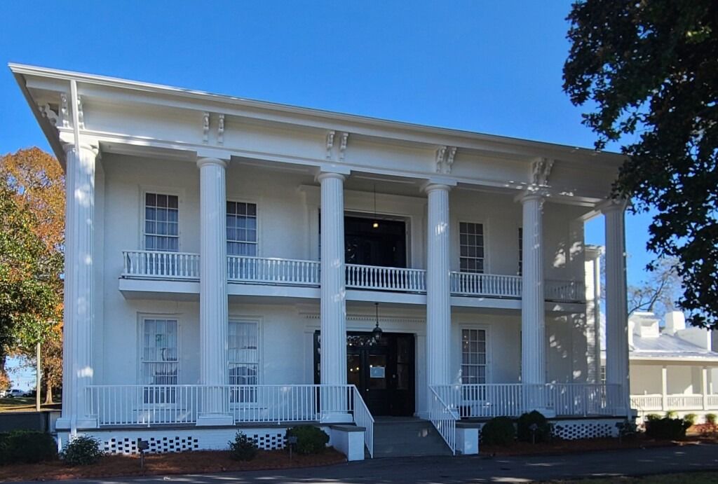 A two-story, white historical mansion with prominent columns, a wide porch, and a symmetric facade against a blue sky.