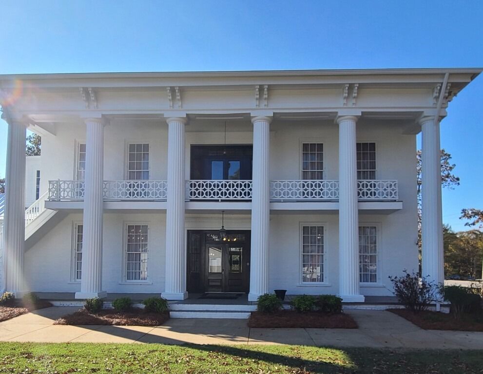 A two-story, white antebellum-style building with a covered porch, six large columns, and an upper balcony.