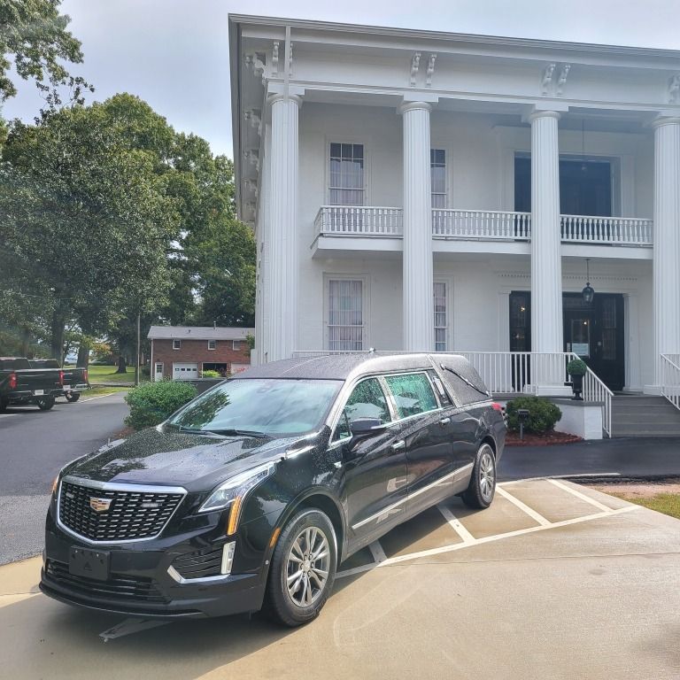A black Cadillac hearse parked on a paved driveway in front of a white, two-story building with large classical columns.
