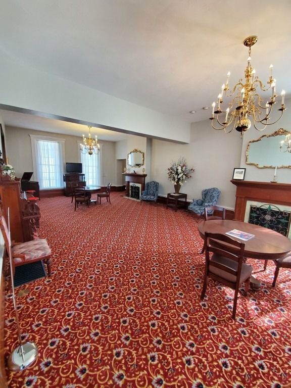 A large, formal room with red patterned carpet, chandeliers, a fireplace, and several seating areas.