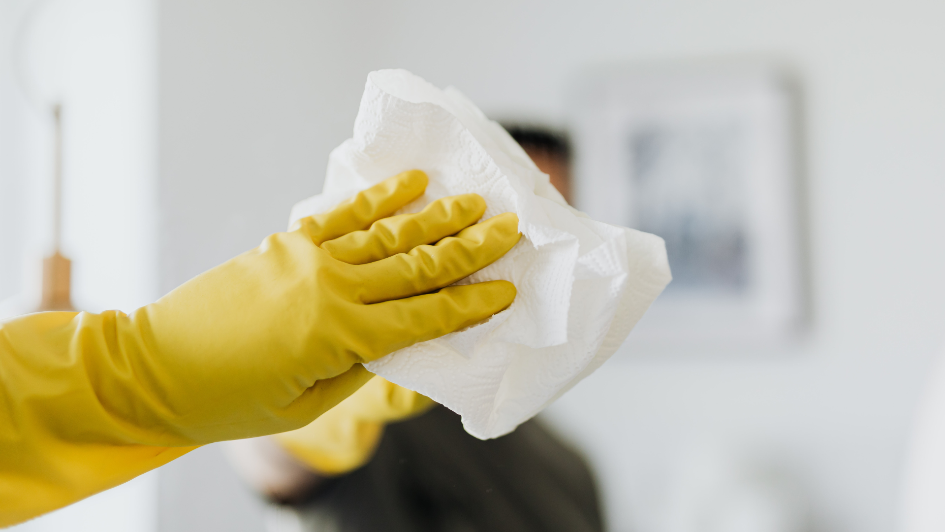 Yellow-gloved hand holding a white cleaning wipe, blurred background with a person and a white wall.