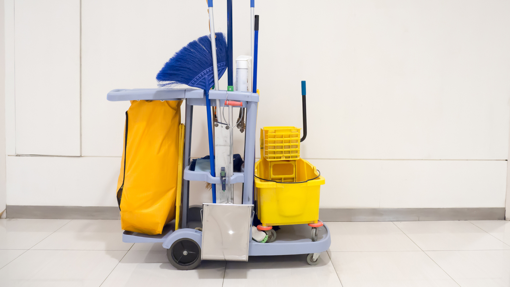 Cleaning cart with yellow bucket, blue brooms, and yellow bag against a white wall.