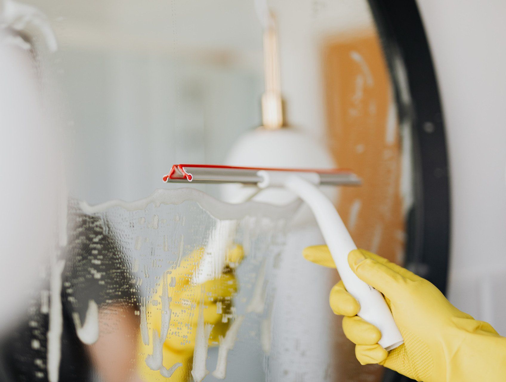 Hands in yellow gloves cleaning a mirror with a squeegee.