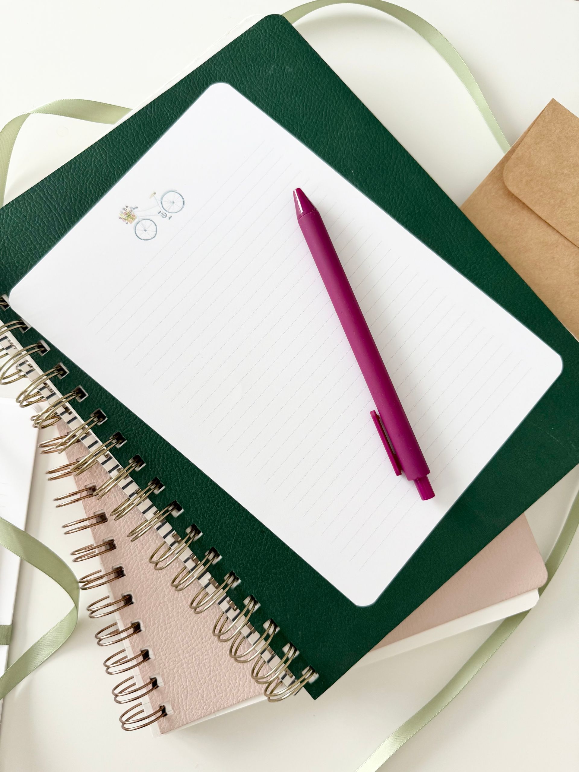 Stationery setup: Green notepad, lined paper with snowman, pink pen, small pink notebook, and a kraft envelope.