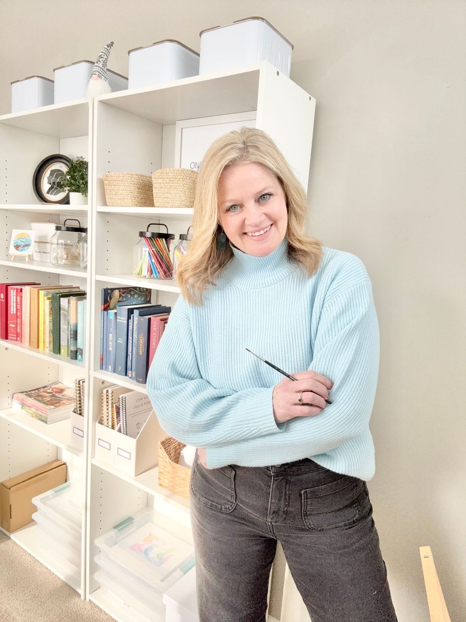 Woman with blonde hair, arms crossed, in front of a desk in a light-filled room.