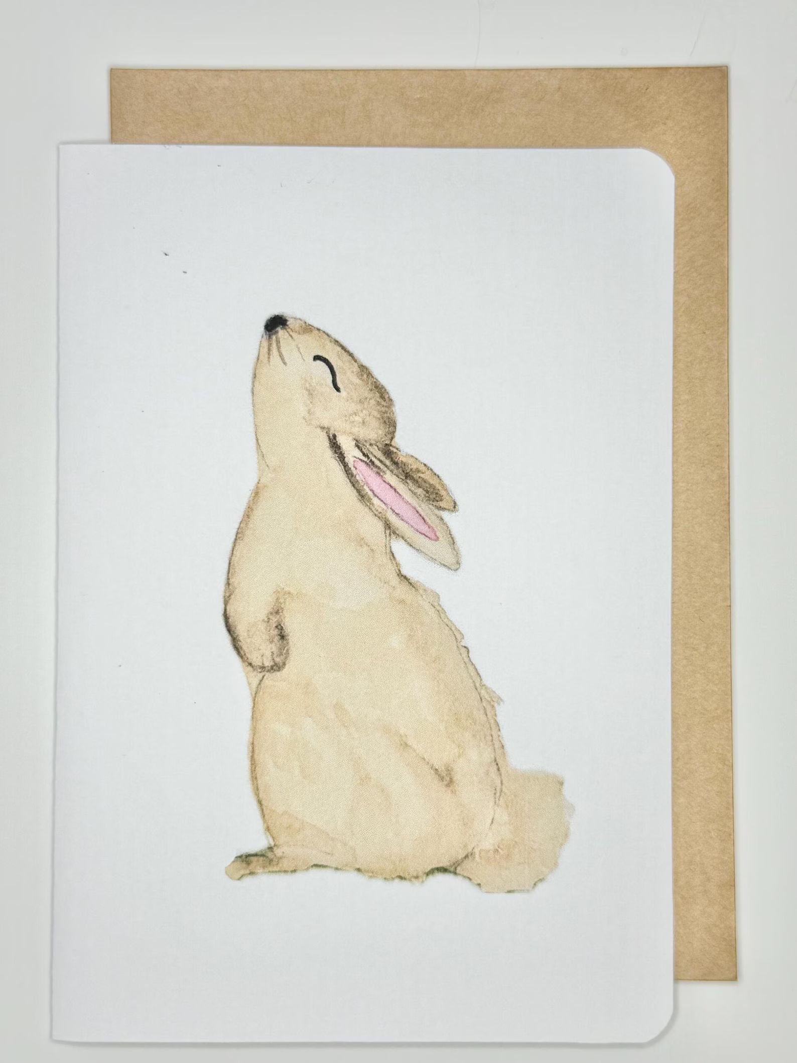 A happy bunny stands on hind legs, looking up. The watercolor drawing is on a white card, with a brown envelope.