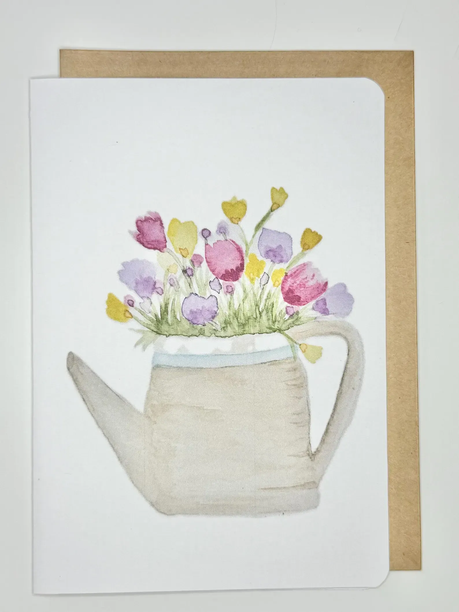 Watercolor of colorful flowers in a watering can on a white card with a brown envelope.