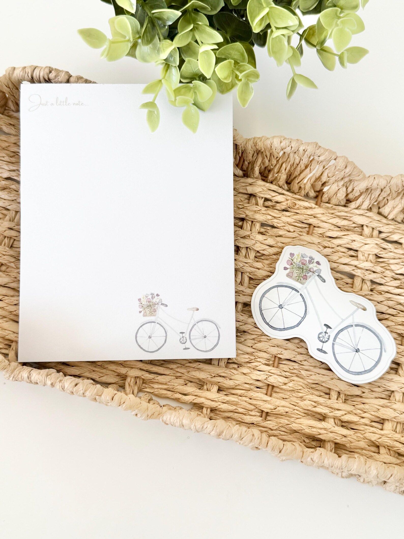 White notepad with bicycle design, next to a matching sticker, on a woven tray with greenery.