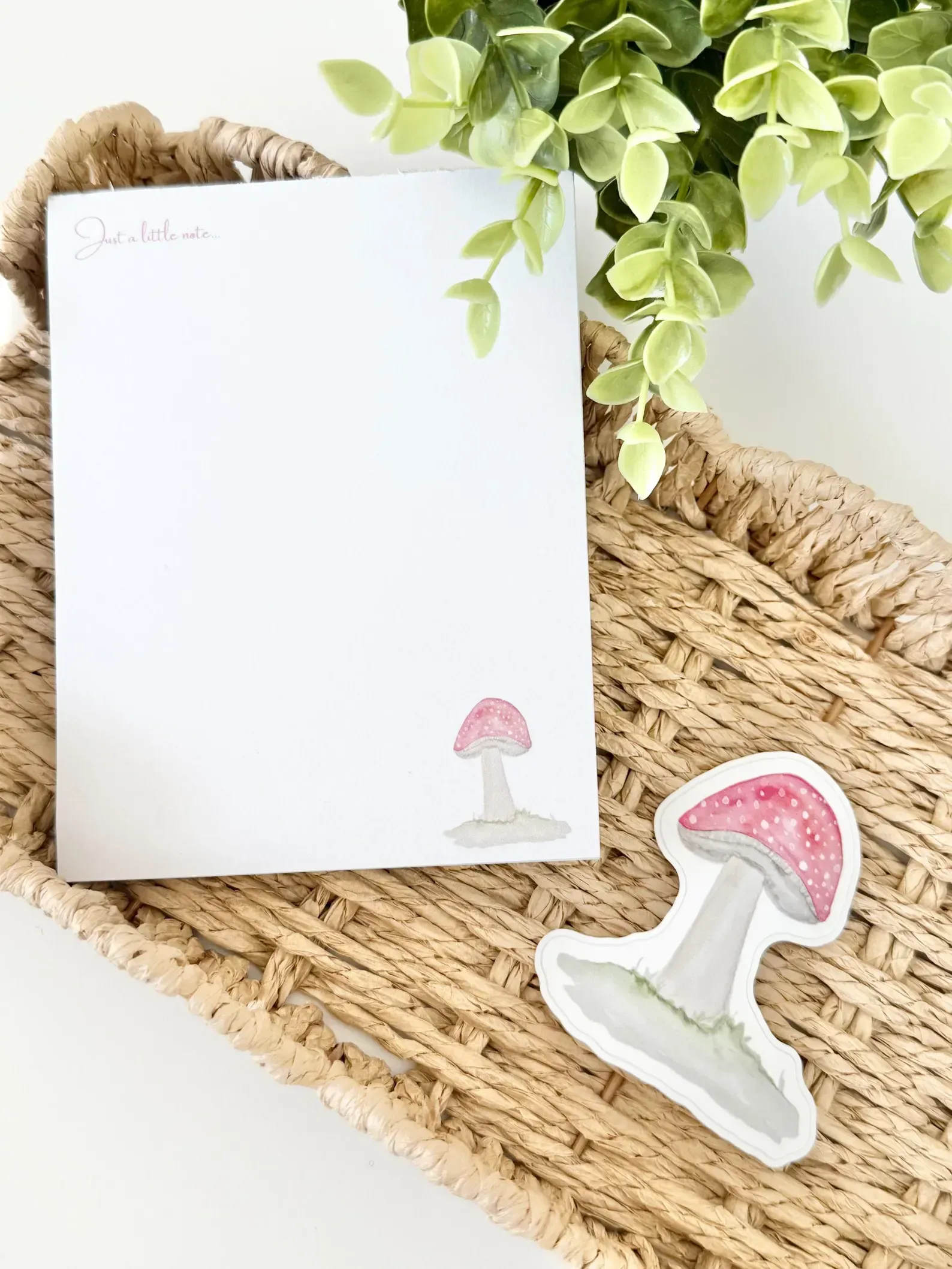 White notepad with mushroom illustration and sticker on woven tray with greenery.