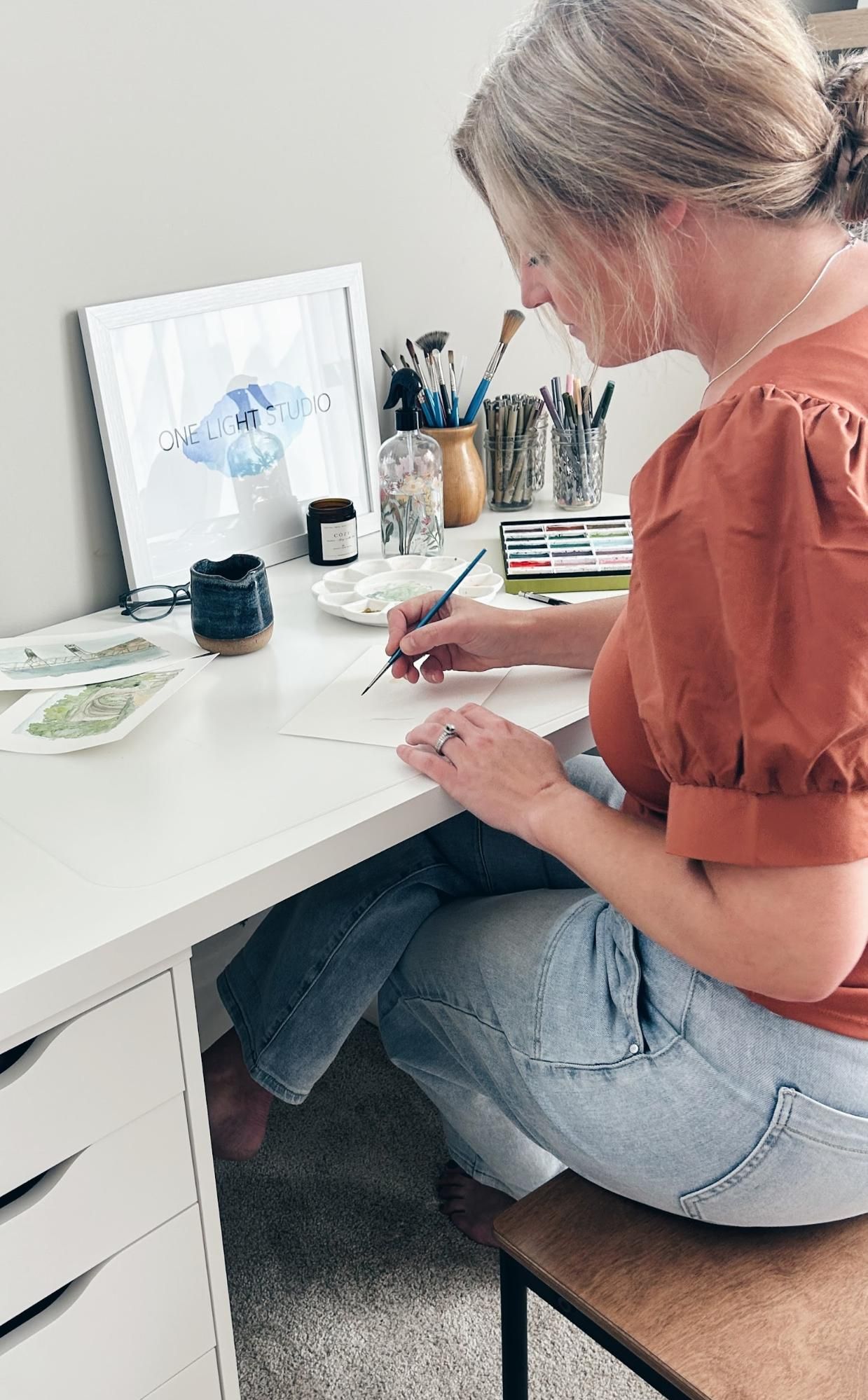 Woman paints watercolor at a white desk, wearing a rust-colored top and jeans, in a light-filled room.