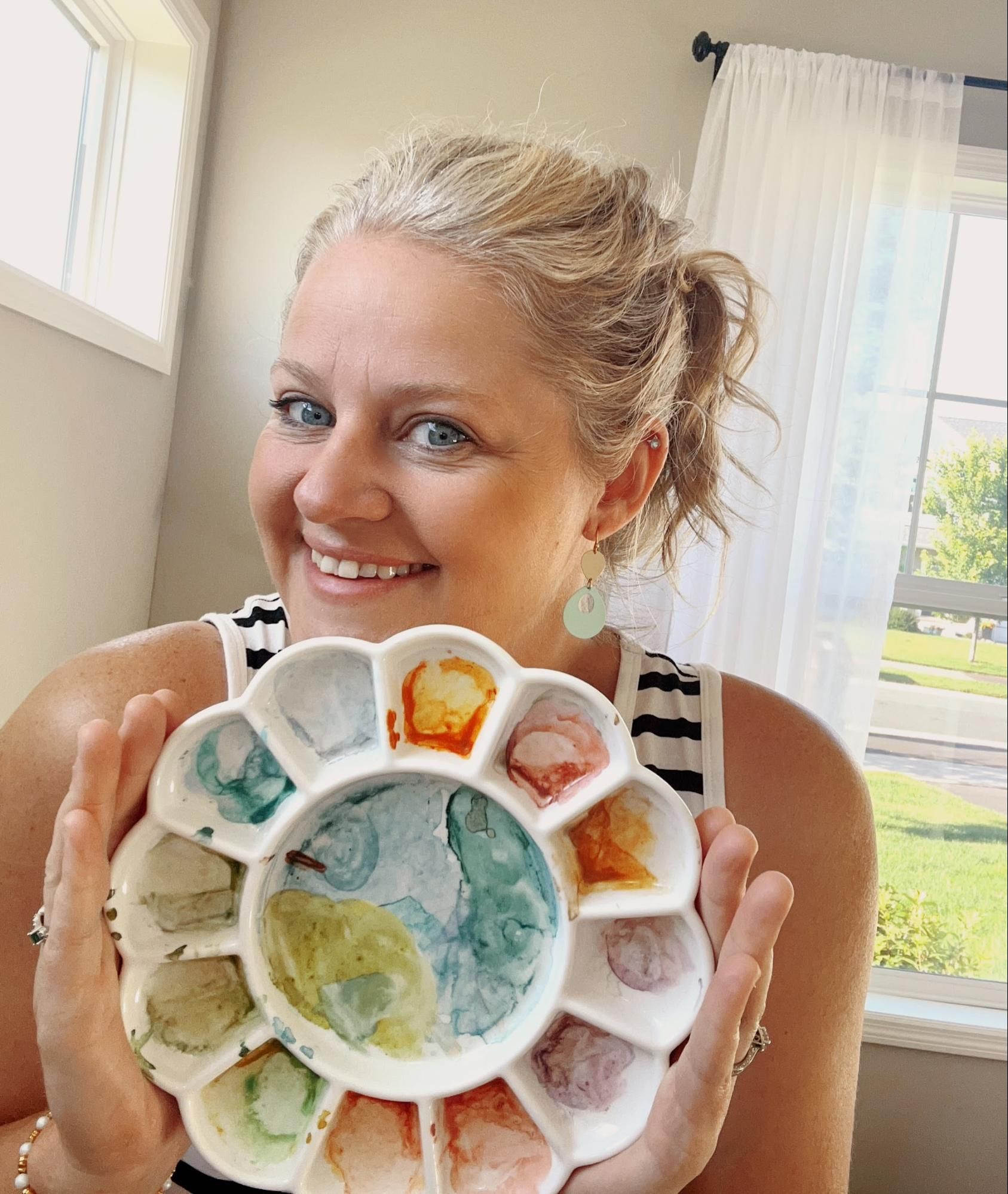 Woman smiling, holding a floral-shaped watercolor palette; setting is indoors with a window.