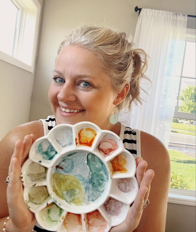 Woman smiling, holding a floral-shaped watercolor palette; setting is indoors with a window.