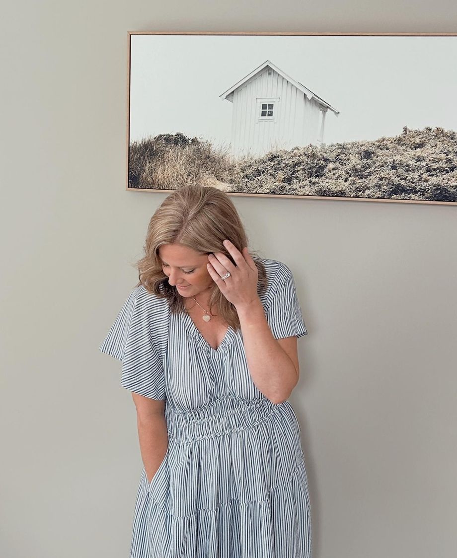 Woman in striped dress, hand on face, stands near a framed picture of a white building on a dune.