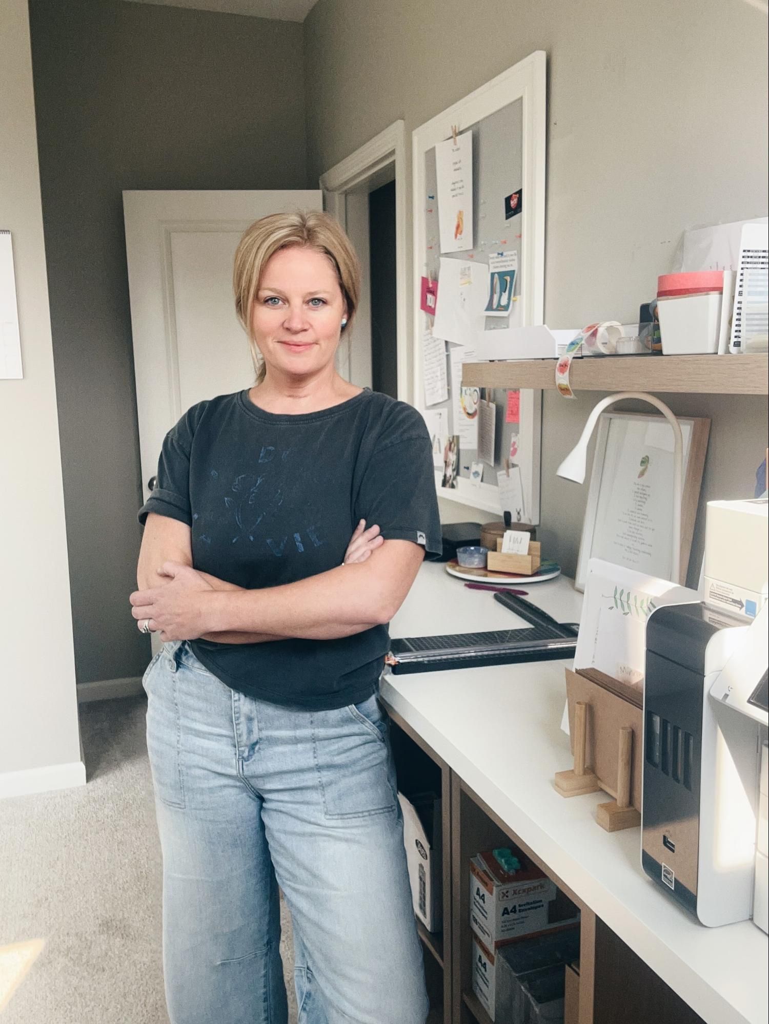 Woman with blonde hair, arms crossed, in front of a desk in a light-filled room.