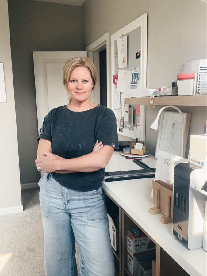 Woman with blonde hair, arms crossed, in front of a desk in a light-filled room.