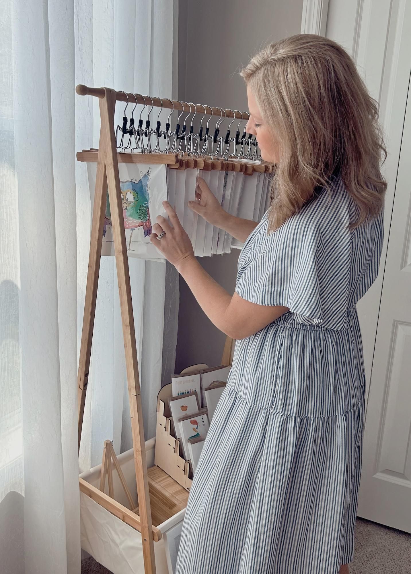 Woman in striped dress arranging artwork on a wooden display rack near a window.