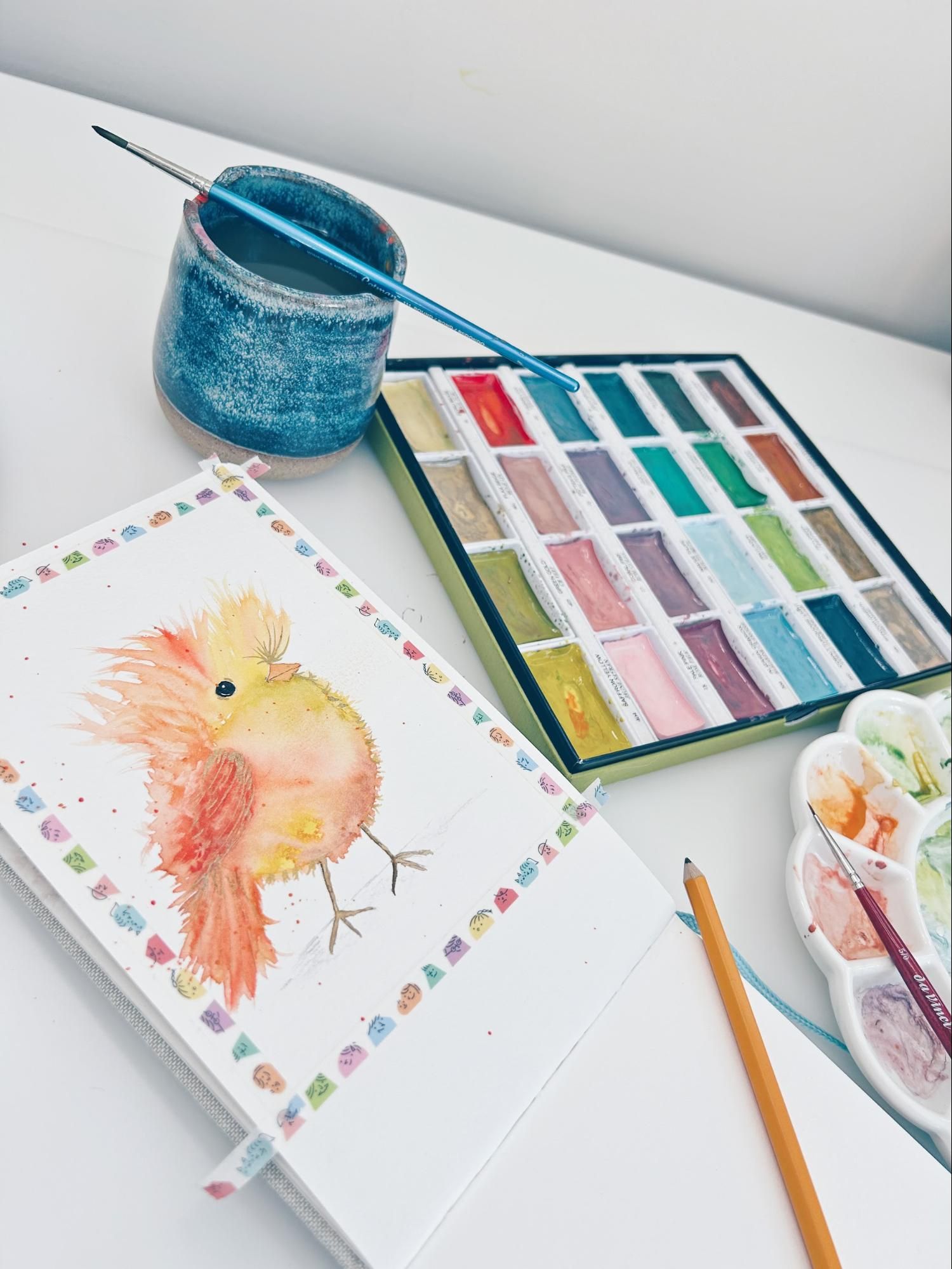 Watercolor bird painting with paints, brush, and water in a white workspace.
