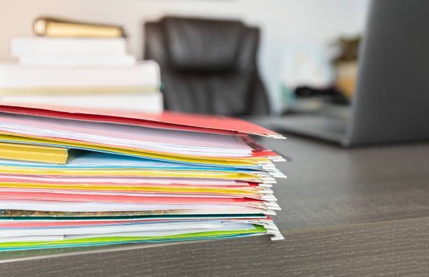 A stack of folders on a desk with a laptop in the background.
