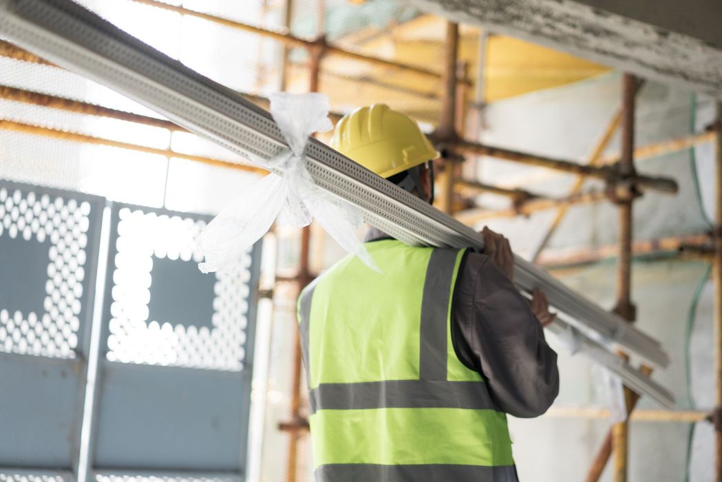 Un operaio edile trasporta lunghe travi di metallo sulle spalle, indossando un casco giallo e un giubbotto di sicurezza.