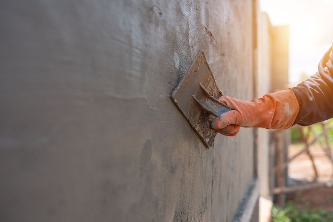 Persona con guanti arancioni che liscia il cemento fresco su un muro con una cazzuola; luce del sole sullo sfondo.