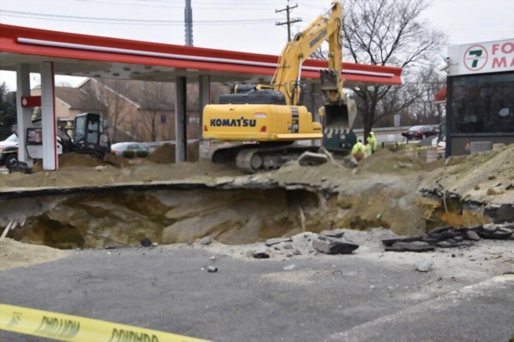Excavator digs at a gas station. Hole in asphalt. 7-Eleven store visible. Construction workers. Yellow caution tape.