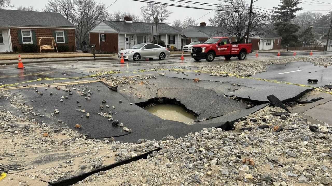 Street collapsed into a large hole; a red truck and a car are nearby, and houses line the street.