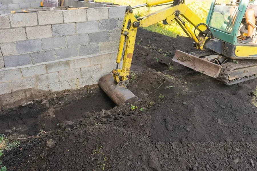 A yellow excavator is digging a hole in the ground next to a brick wall for sinkhole repair