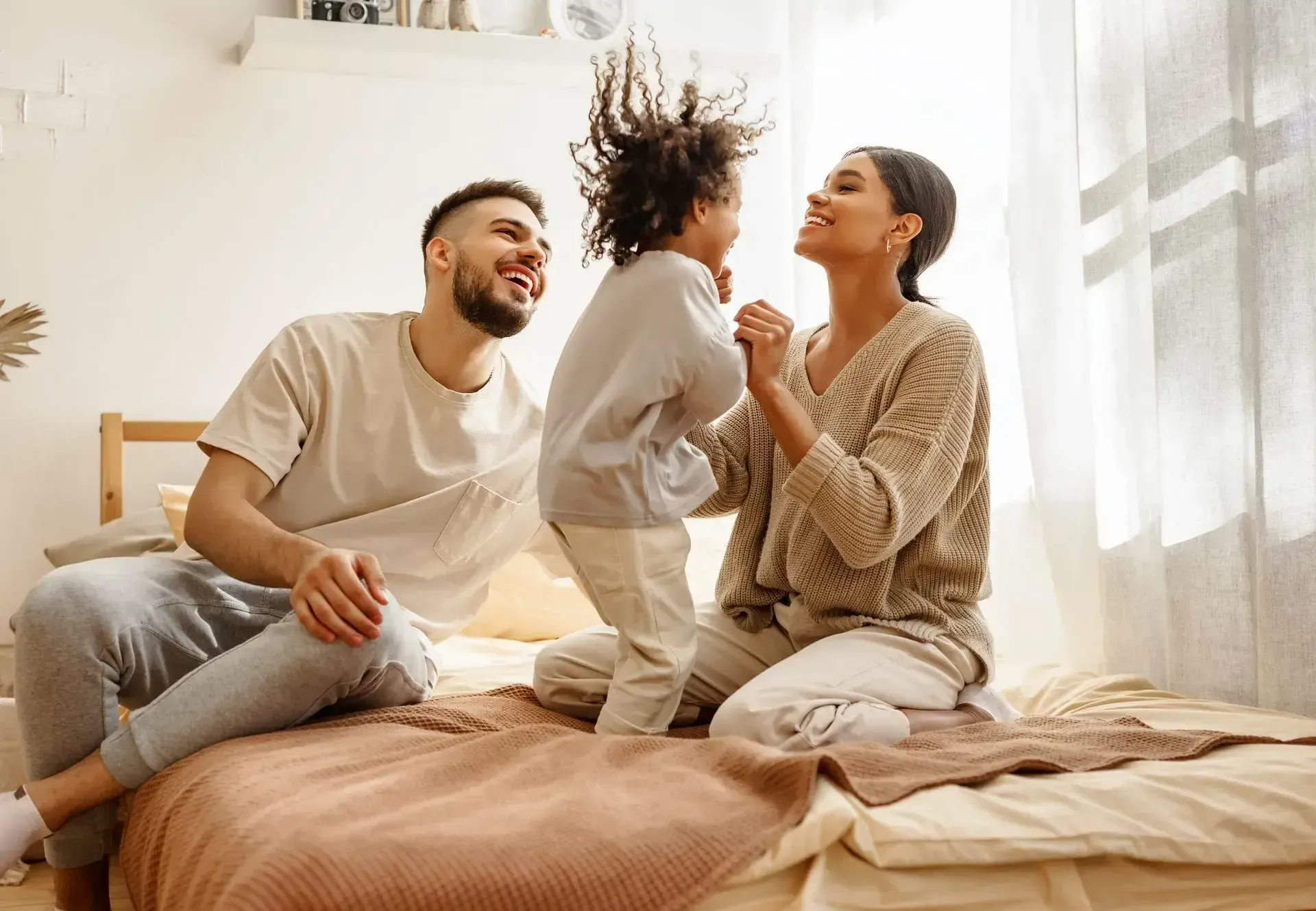 A family is sitting on a bed with a child jumping in the air.