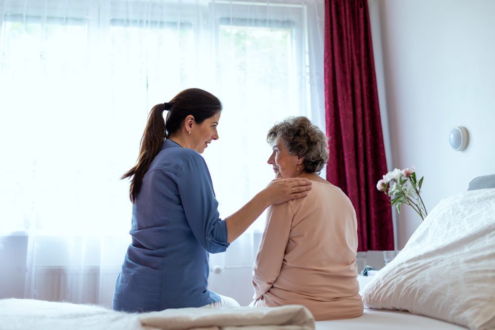 Caregiver in Blue Shirt Comforts Elderly Woman on Bed, Hand on Her Shoulder — Elder Care Connect in Highland Park, QLD