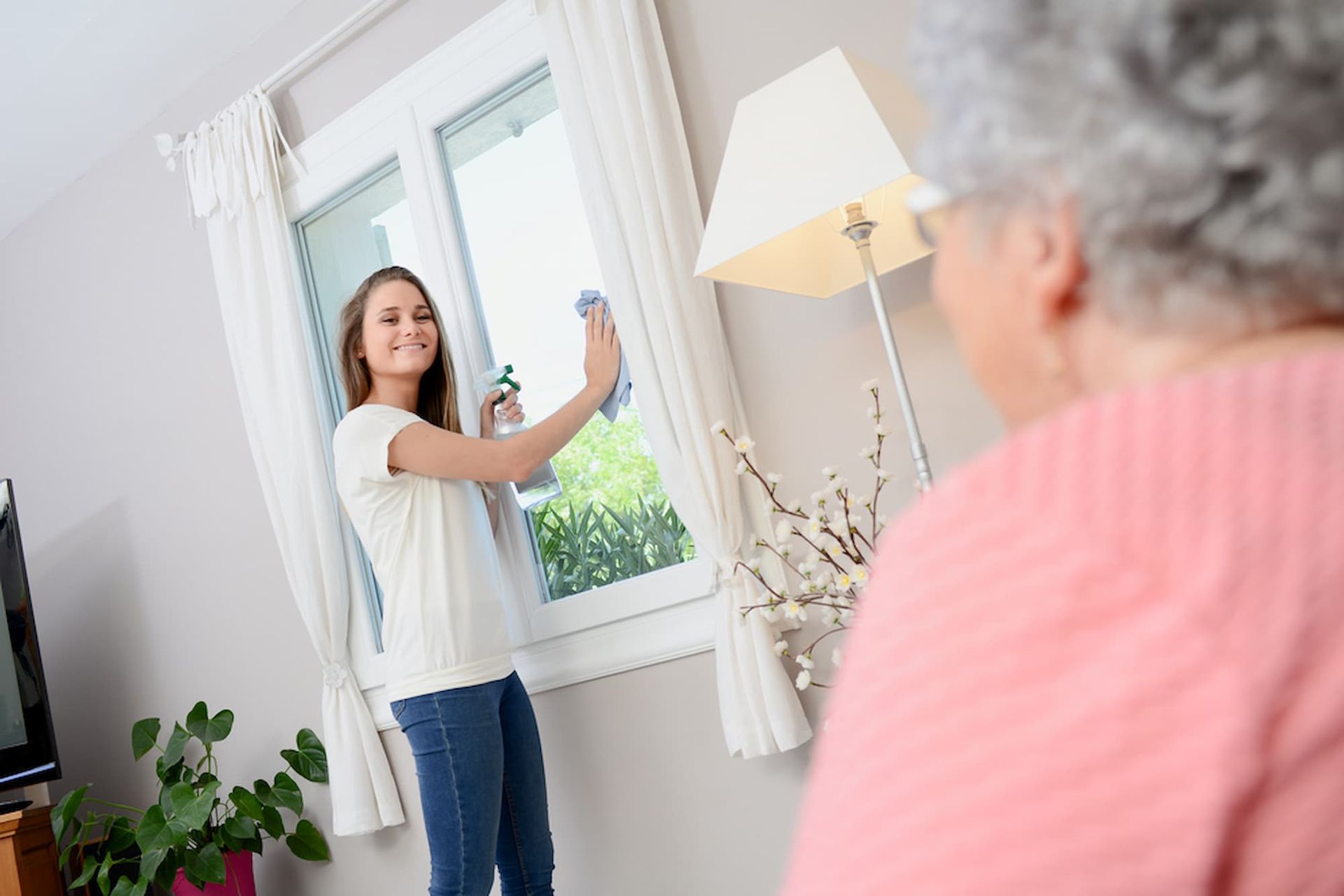 Young Woman Cleaning a Window, Older Woman Watches in A Bright Living Room — Elder Care Connect in Palm Beach, QLD