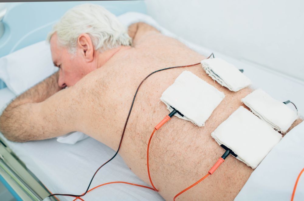 An elderly man is laying on a bed with electrodes on his back.