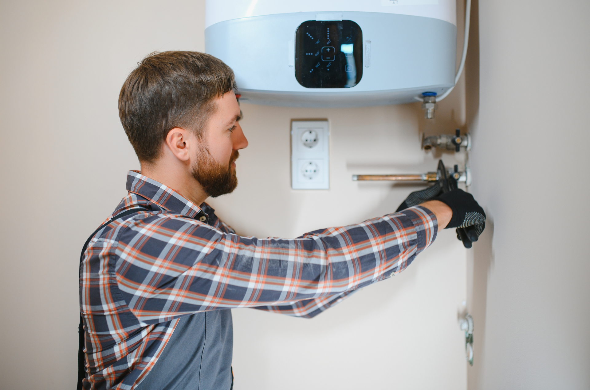 Plumber adjusting a pipe on a water heater. He wears gloves and a plaid shirt, working in a light-colored room.
