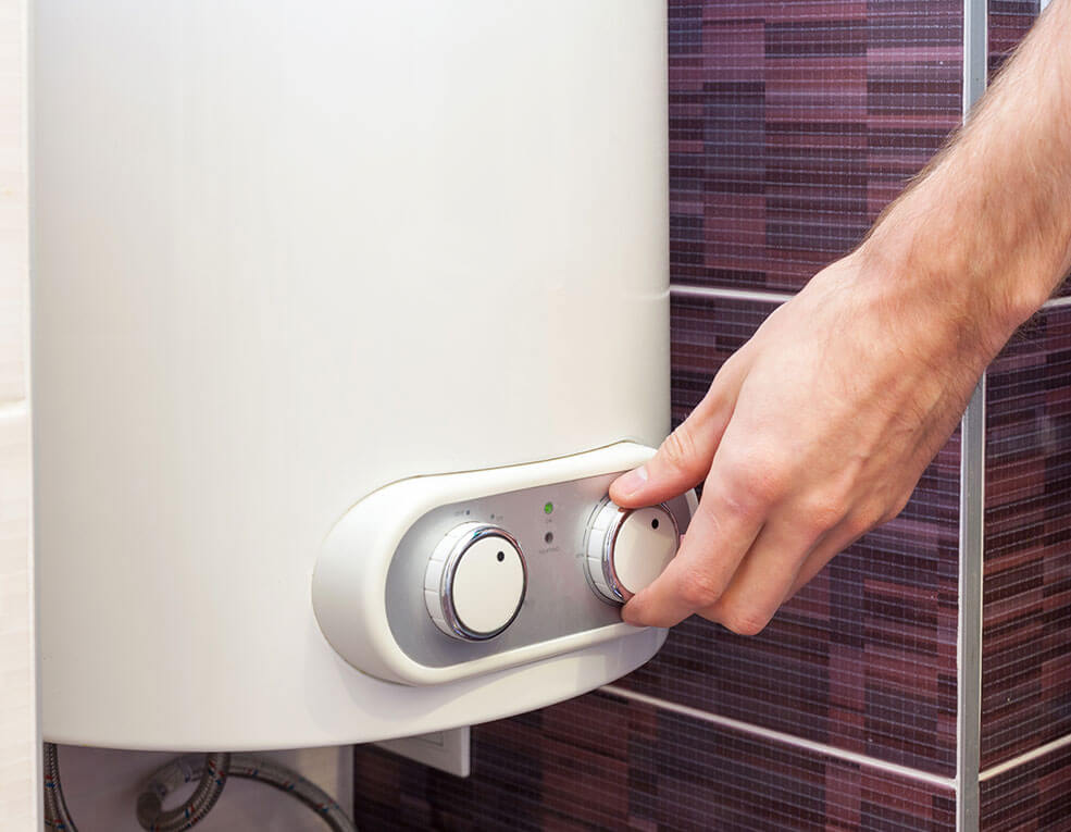Hand turning a dial on a white water heater mounted on a tile wall.