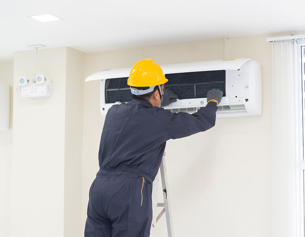 HVAC technician in yellow hard hat repairs a wall-mounted air conditioner unit indoors.