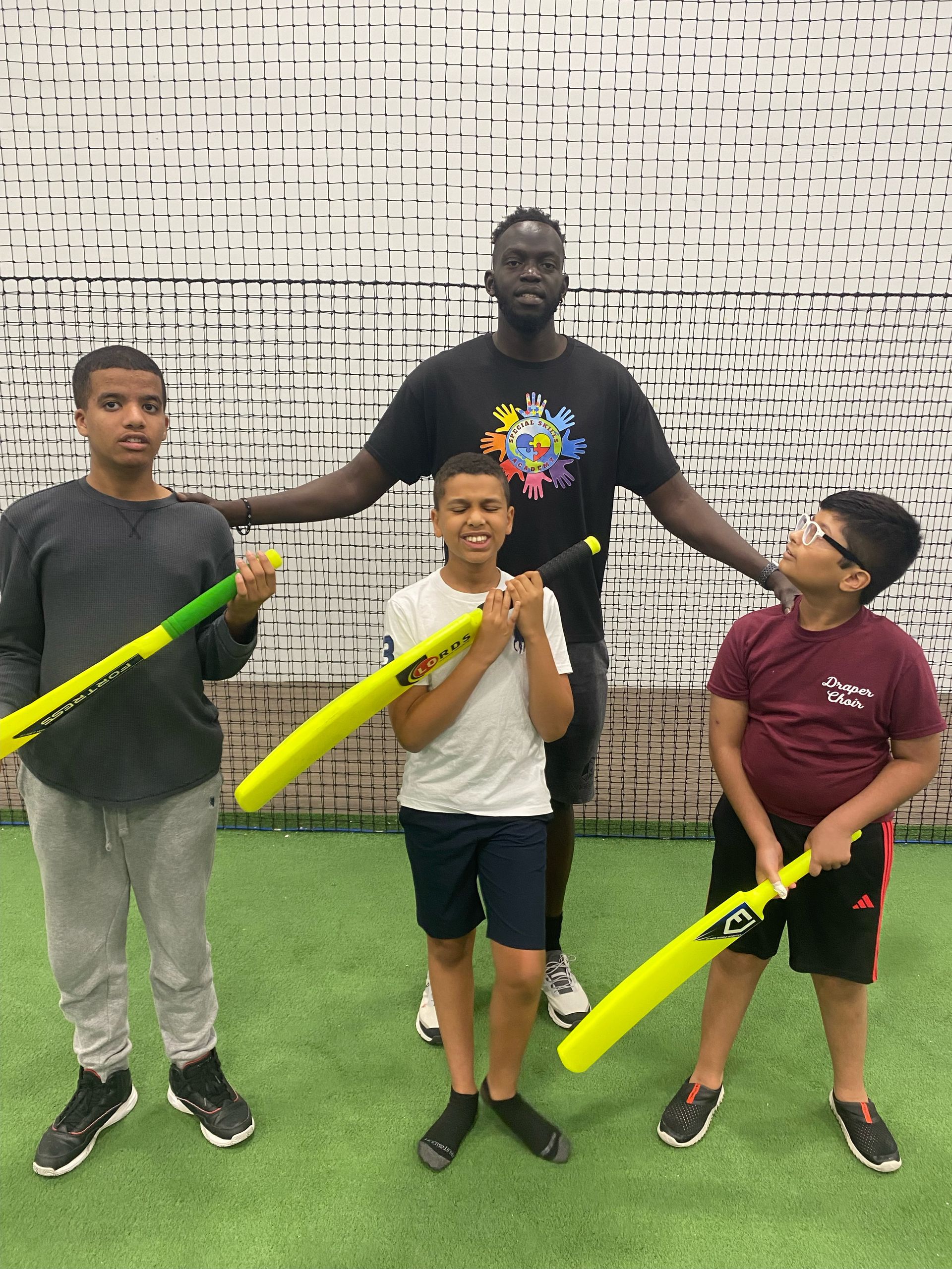 Group of kids and tall man holding yellow bats, posing on green turf; neutral background.