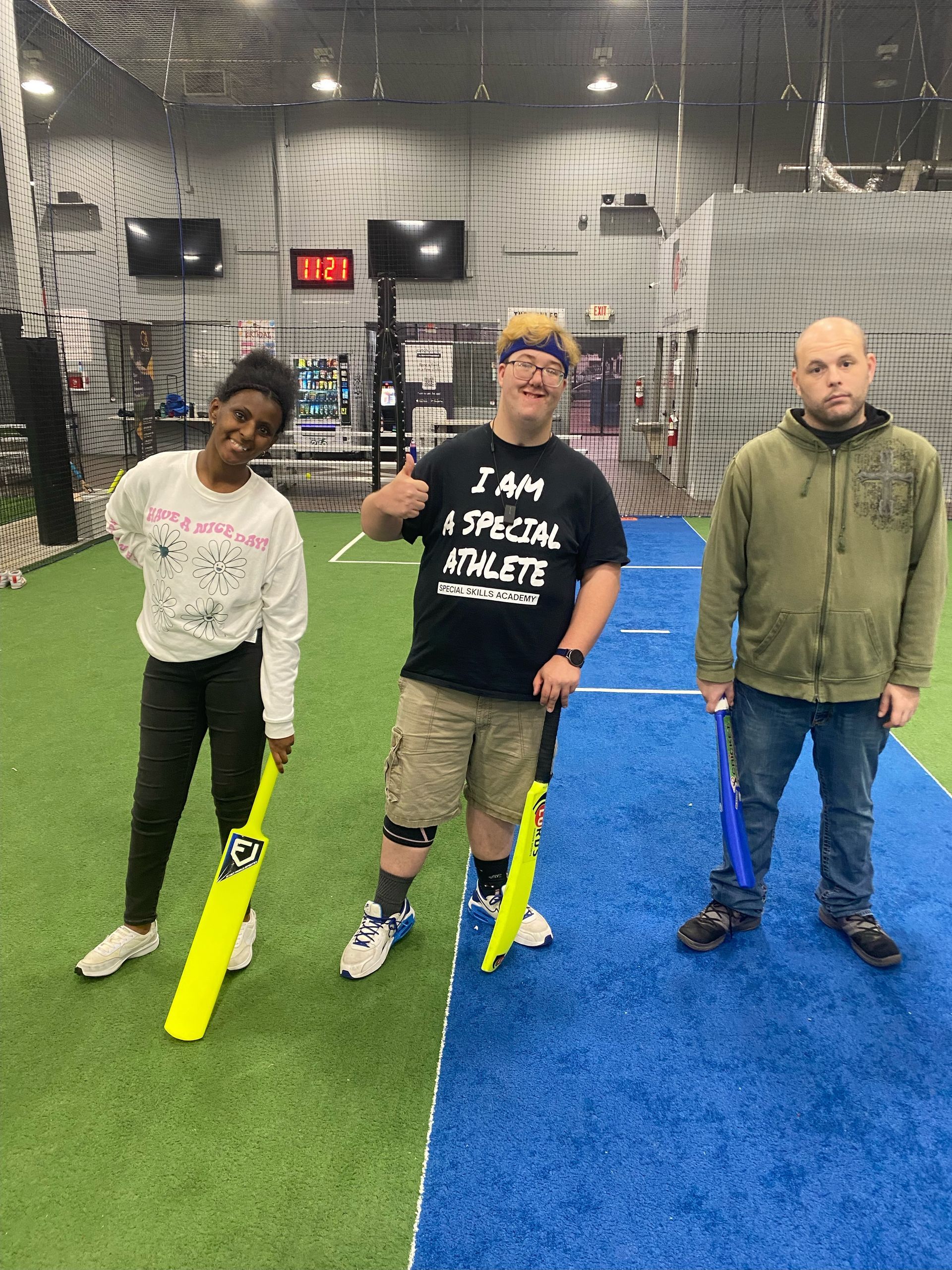 Three people with cricket bats on a blue/green turf. One gives a thumbs up. Indoors, netting in background.