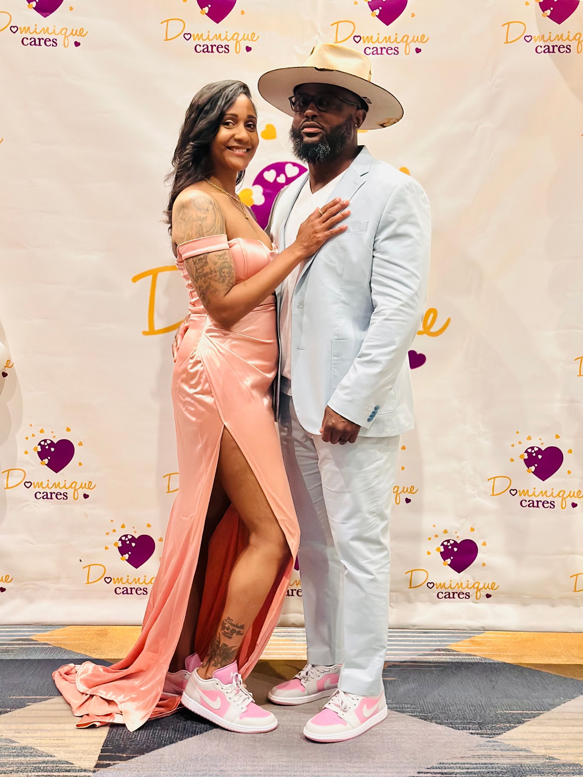 Couple poses in front of a backdrop. Woman in peach gown with high slit, man in light blue suit, both wear pink sneakers.