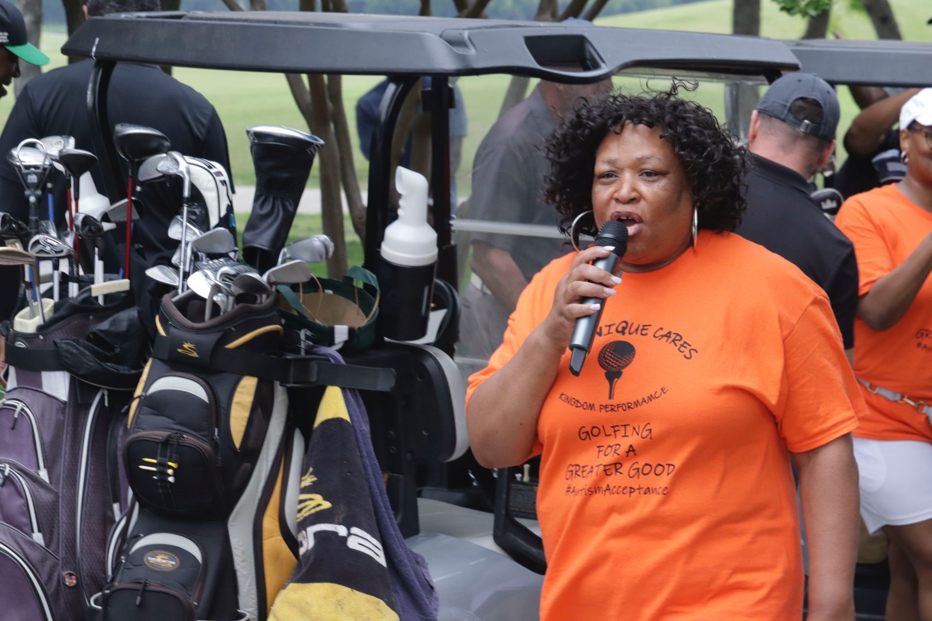 Woman with microphone speaking at golf event. She wears an orange shirt, next to golf bags.